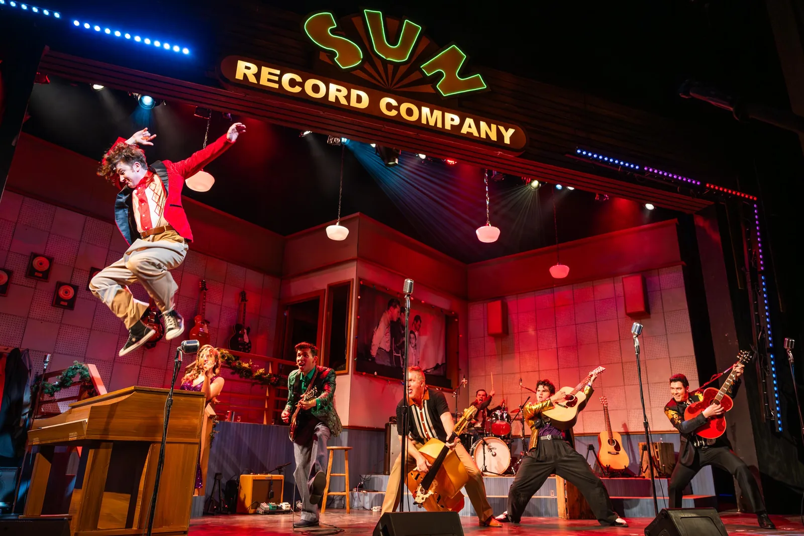 A performer leaps mid-air in front of a band on the Sun Record Company set, bathed in red stage light, in a production of Million Dollar Quartet.