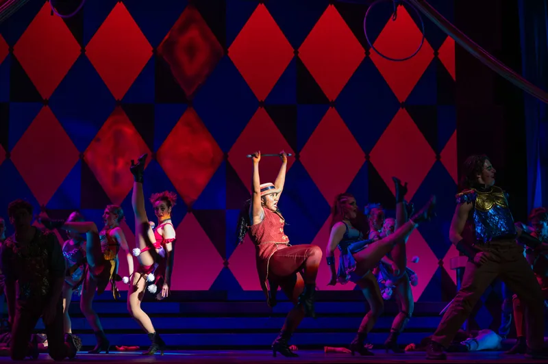 A dancer hangs from an overhead bar as the ensemble kicks in front of a wall of red and blue diamonds.