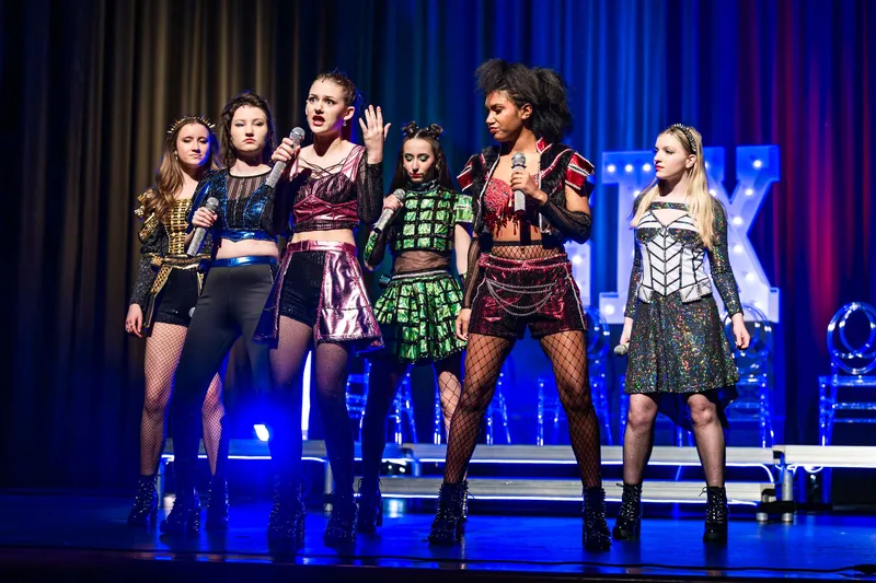 Six performers in sequined, studded costumes sing into handheld microphones under blue concert lighting.