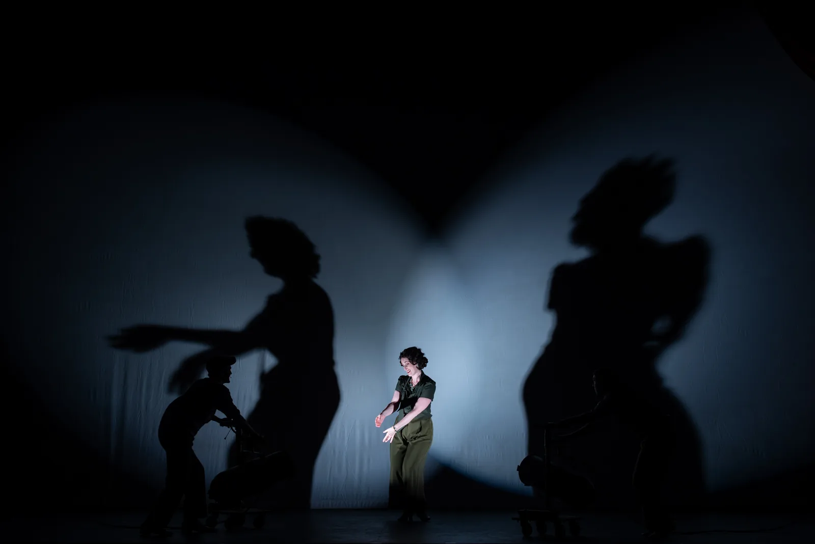 A lone actor stands in a pool of white light while huge shadows of other performers stretch across the back wall.