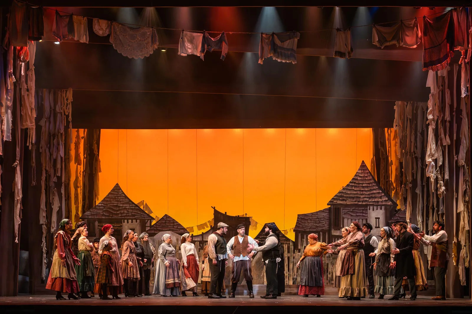 Villagers in period dress fill the stage before a glowing orange sky and a village of rooftops, in a production of Fiddler on the Roof.