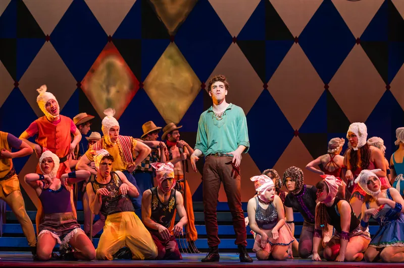 An ensemble in patchwork costumes crouches around a young man in a green shirt against a harlequin-diamond backdrop.