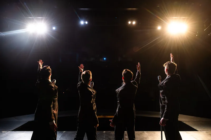 Four performers in dark suits, seen from behind, raise their arms toward blazing stage lights.