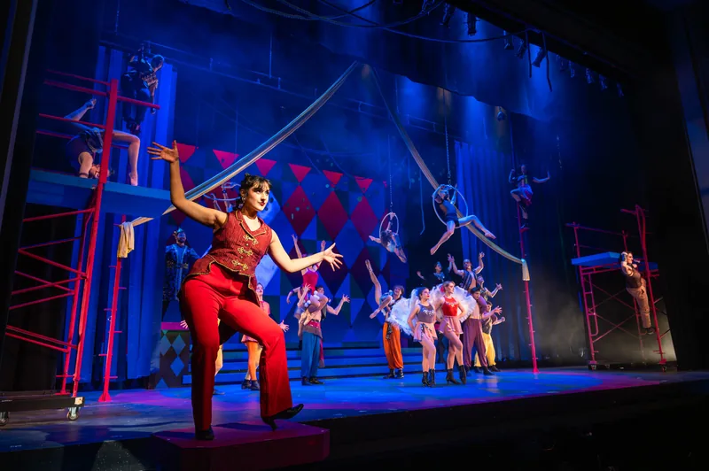 A dancer in a red jumpsuit strikes a pose as aerialists and the ensemble perform on a blue-lit circus set with red scaffolding.