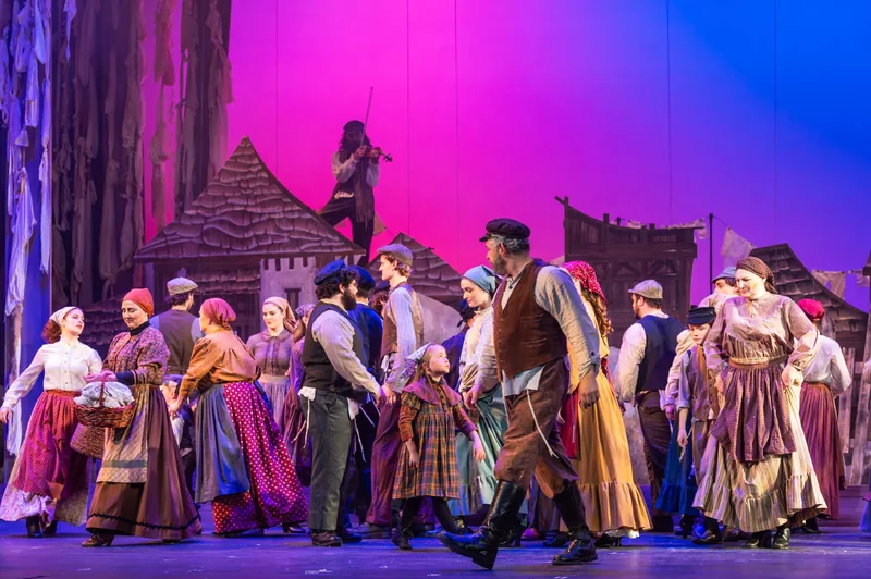 Villagers in period costume dance across the stage under magenta light while a fiddler plays on a rooftop above, in a production of Fiddler on the Roof.