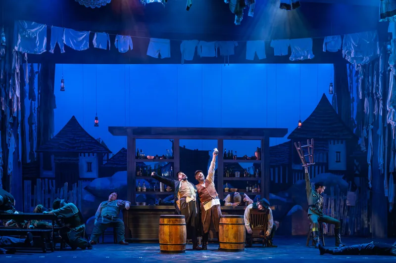 Townspeople gather in a tavern scene under deep blue light, with laundry strung above the village set.