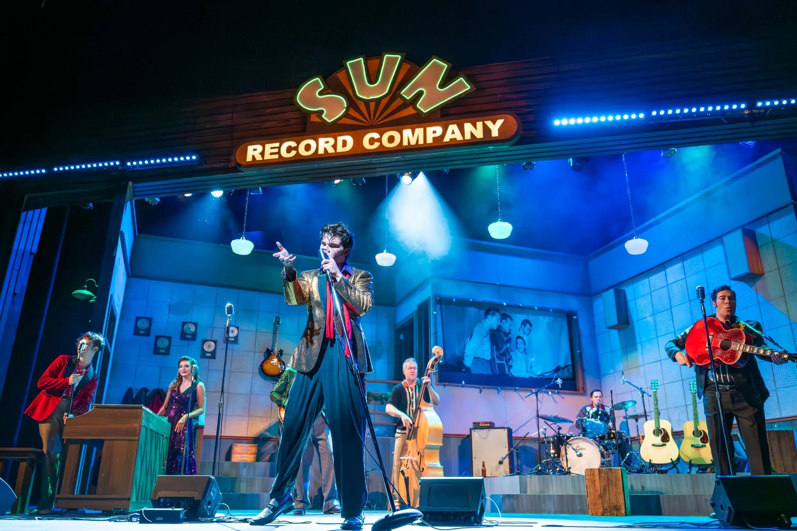 A singer in a gold jacket points toward the audience from the center of the band on the Sun Record Company set, lit in blue.