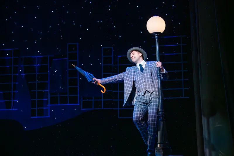 A performer in a plaid suit swings from a lamppost with an umbrella against a starry night backdrop.