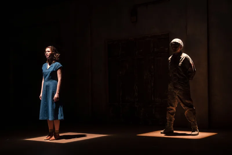 Two actors stand apart in separate shafts of light on a dark, sparse stage.