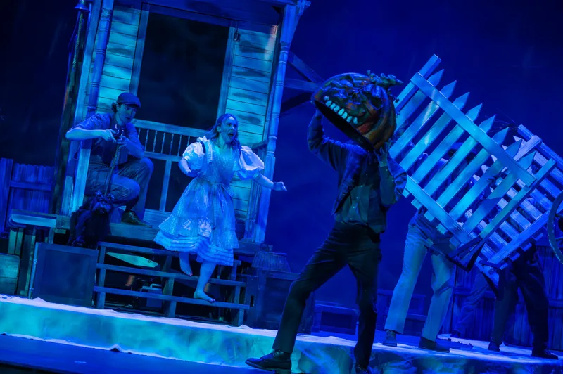 Under blue light, an actor carries a giant plant puppet past a run-down storefront while a woman in a white dress looks on.