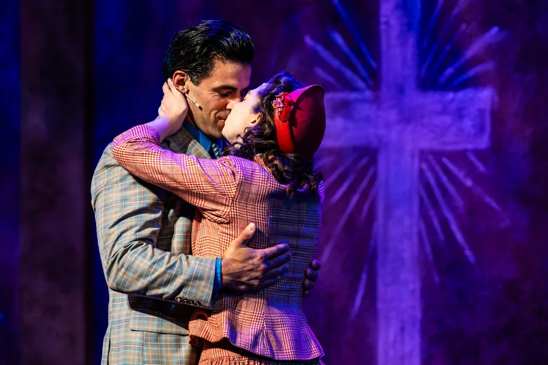 Two actors share a kiss in front of a glowing cross on the wall behind them.
