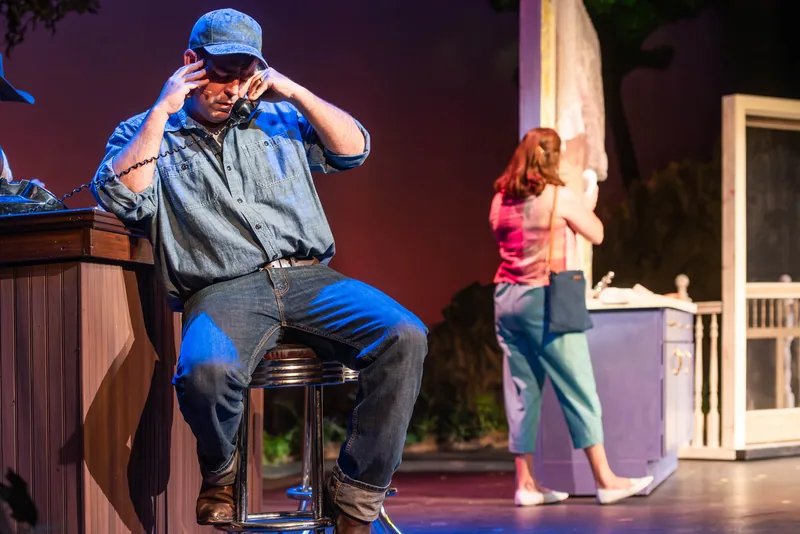 A man in a denim shirt and cap talks on a corded phone while a woman in pink walks away across a small-town set.