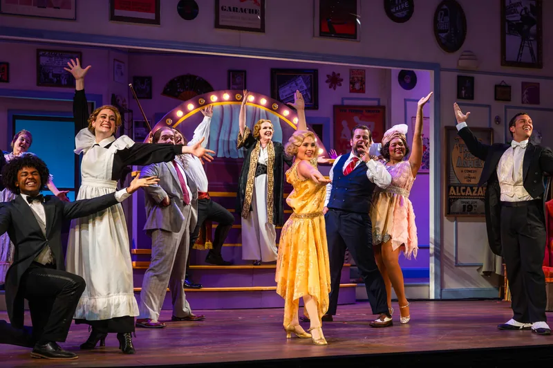 The ensemble celebrates with arms raised on a colorful 1920s theatre-lobby set.