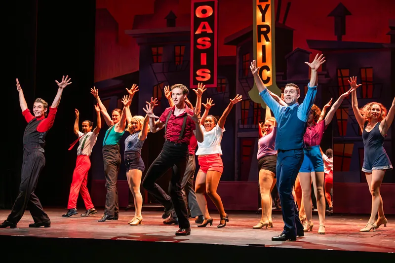 Dancers in bright costumes throw their arms up in front of neon theatre signs reading Oasis and Lyric.