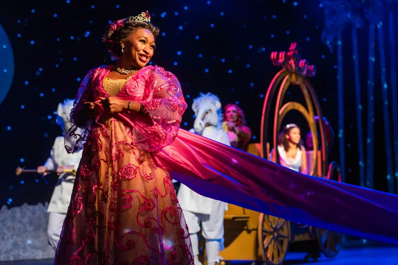 A fairy godmother in a flowing pink gown smiles beside a golden carriage under a starry sky.