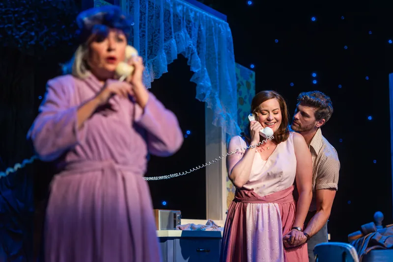 A woman in a bathrobe talks on a telephone in the foreground while a couple shares the call behind her.