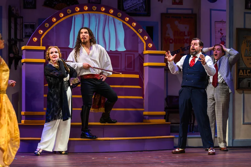 Actors react dramatically on a vaudeville-style stage framed by marquee lights.