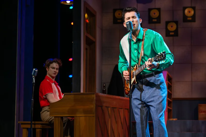 A singer in a green shirt plays electric guitar at a vintage microphone while a pianist looks on.