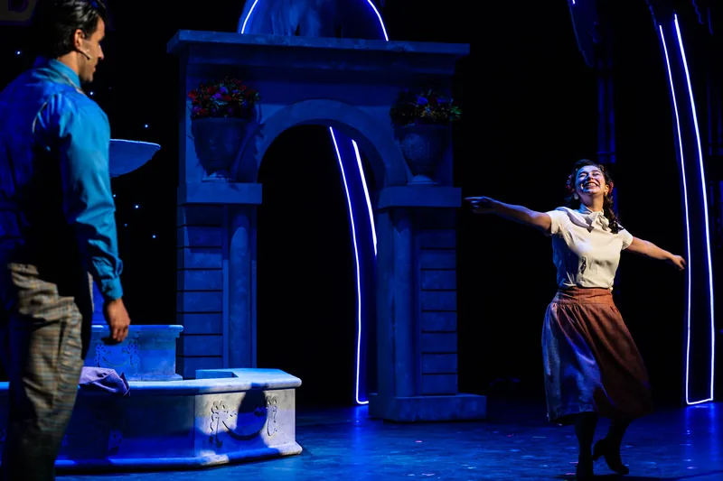 A young woman in a peasant dress runs with arms outstretched past a stone fountain lit in blue.